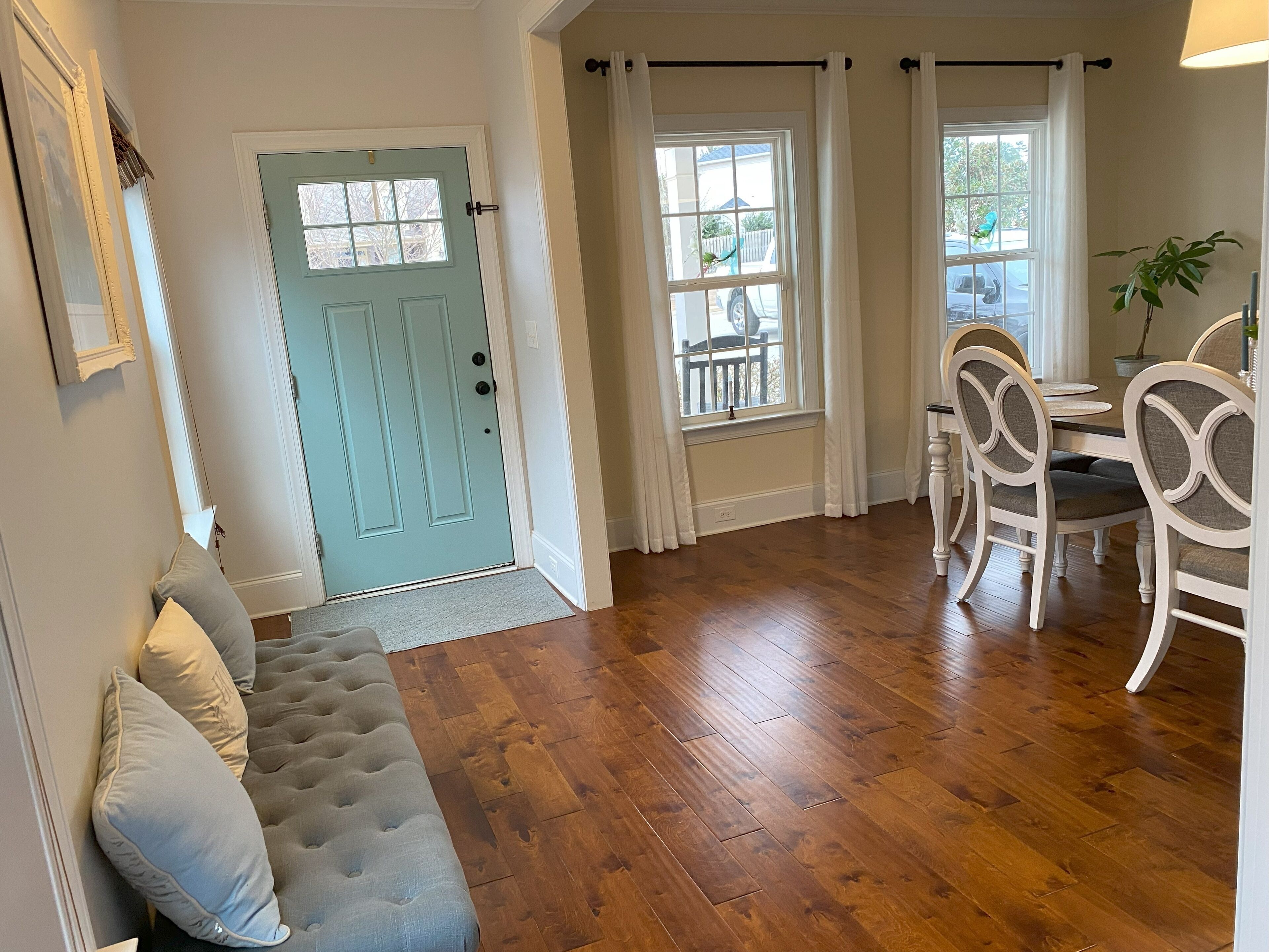 A room with a light blue door, a wooden floor, a dining area with white chairs, and a gray sofa with cushions.