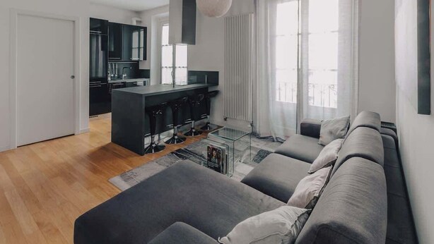 Una sala de estar moderna con un sofá gris, una mesa de centro de vidrio y una cocina al fondo.