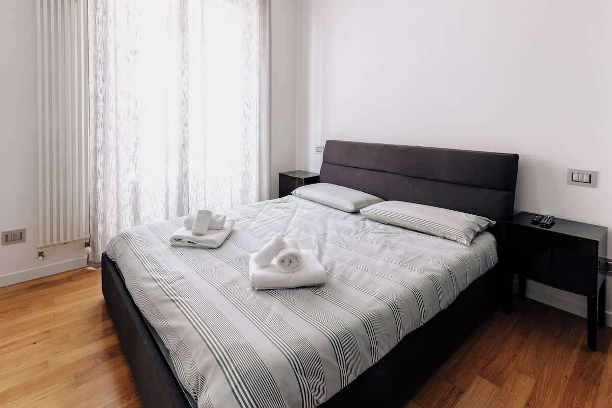 A neatly made bed with a striped bedspread and pillows, a bedside table with a remote control, and a window with sheer curtains.