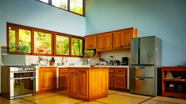 Une cuisine dotée de meubles en bois, d’un îlot central, d’appareils électroménagers en acier inoxydable et de grandes fenêtres offrant une vue sur la verdure.