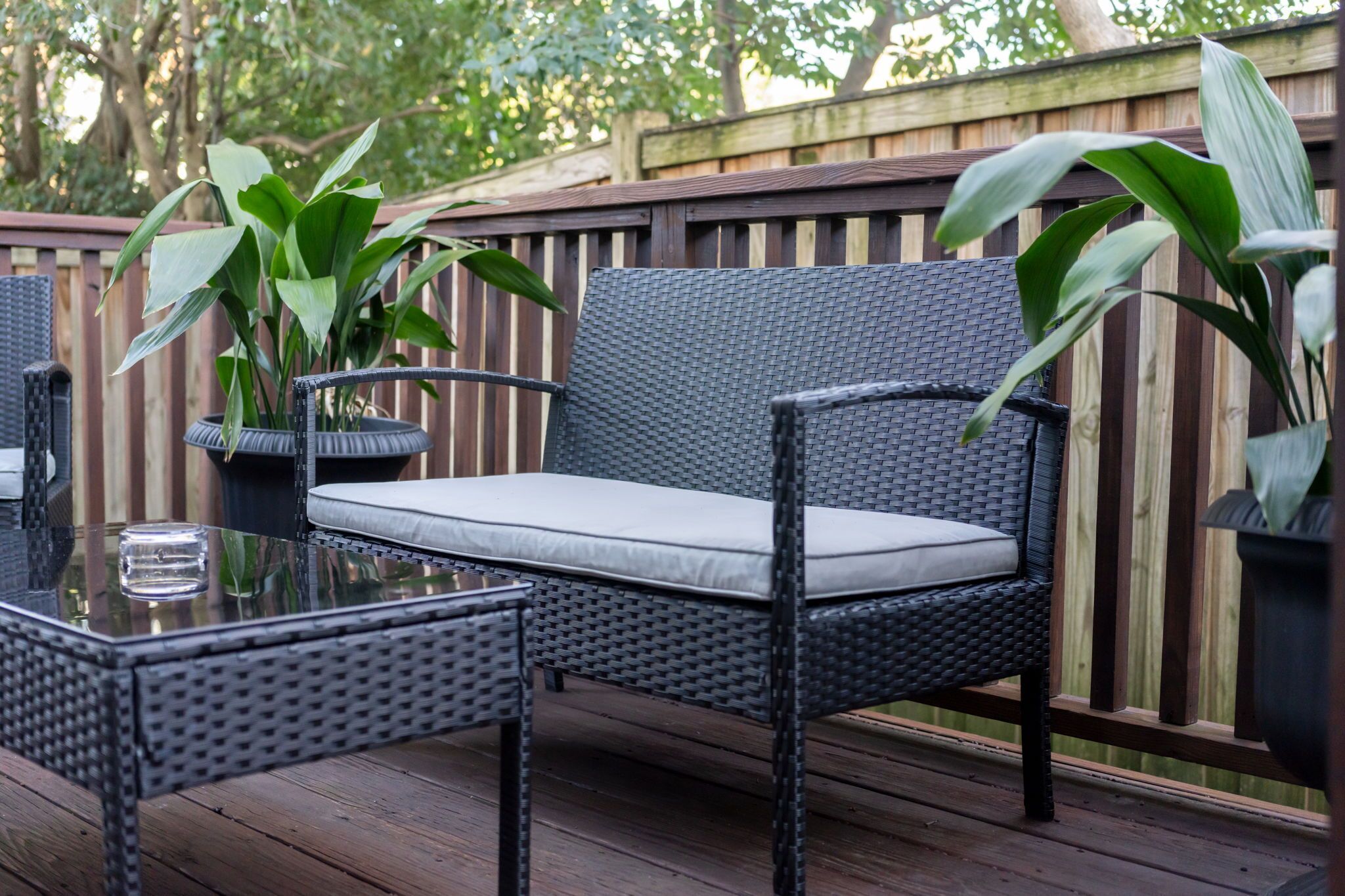Un espacio de estar al aire libre con muebles de mimbre, un banco acolchado y una mesa con superficie de vidrio, rodeado de plantas en macetas.