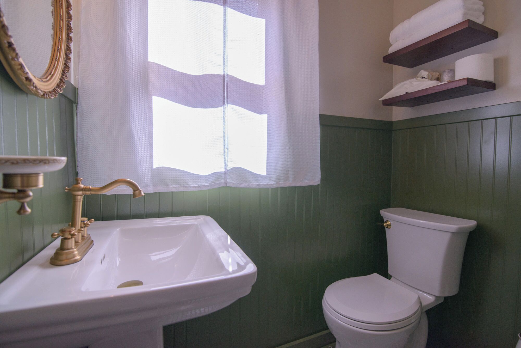 Une salle de bain avec un lavabo blanc, des toilettes et une fenêtre avec un rideau blanc.