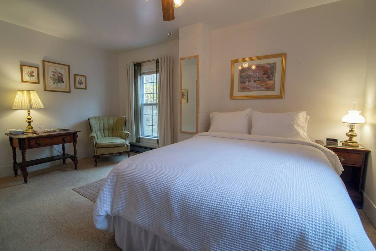 A neatly arranged bedroom with a large bed, a chair, a desk, and a window with curtains.