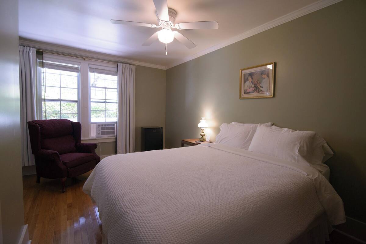 A bedroom with a large bed, a chair, a ceiling fan, a nightstand with a lamp, and a window with curtains.