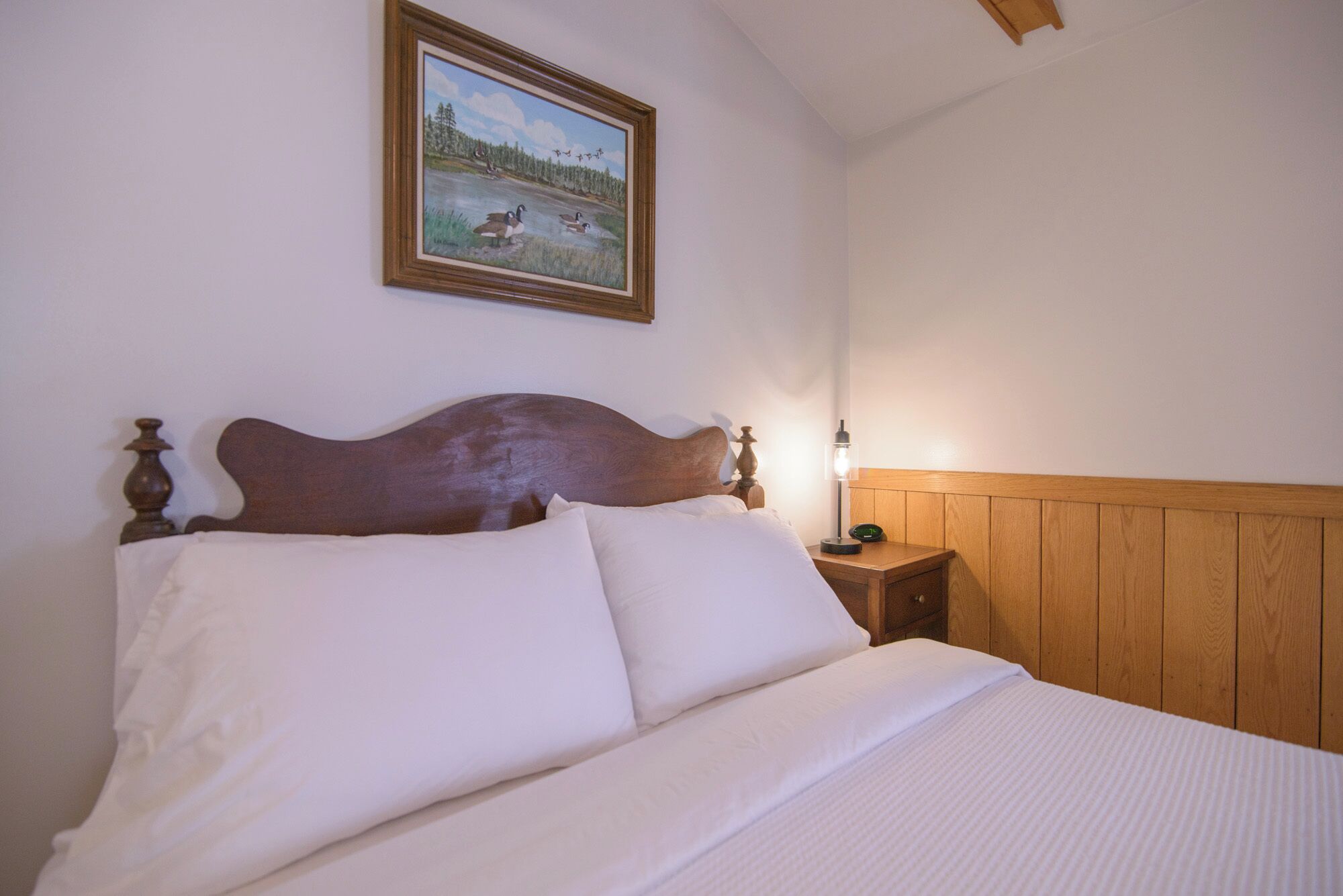 A neatly made bed with white linens, a wooden headboard, and a framed painting of birds on the wall above.