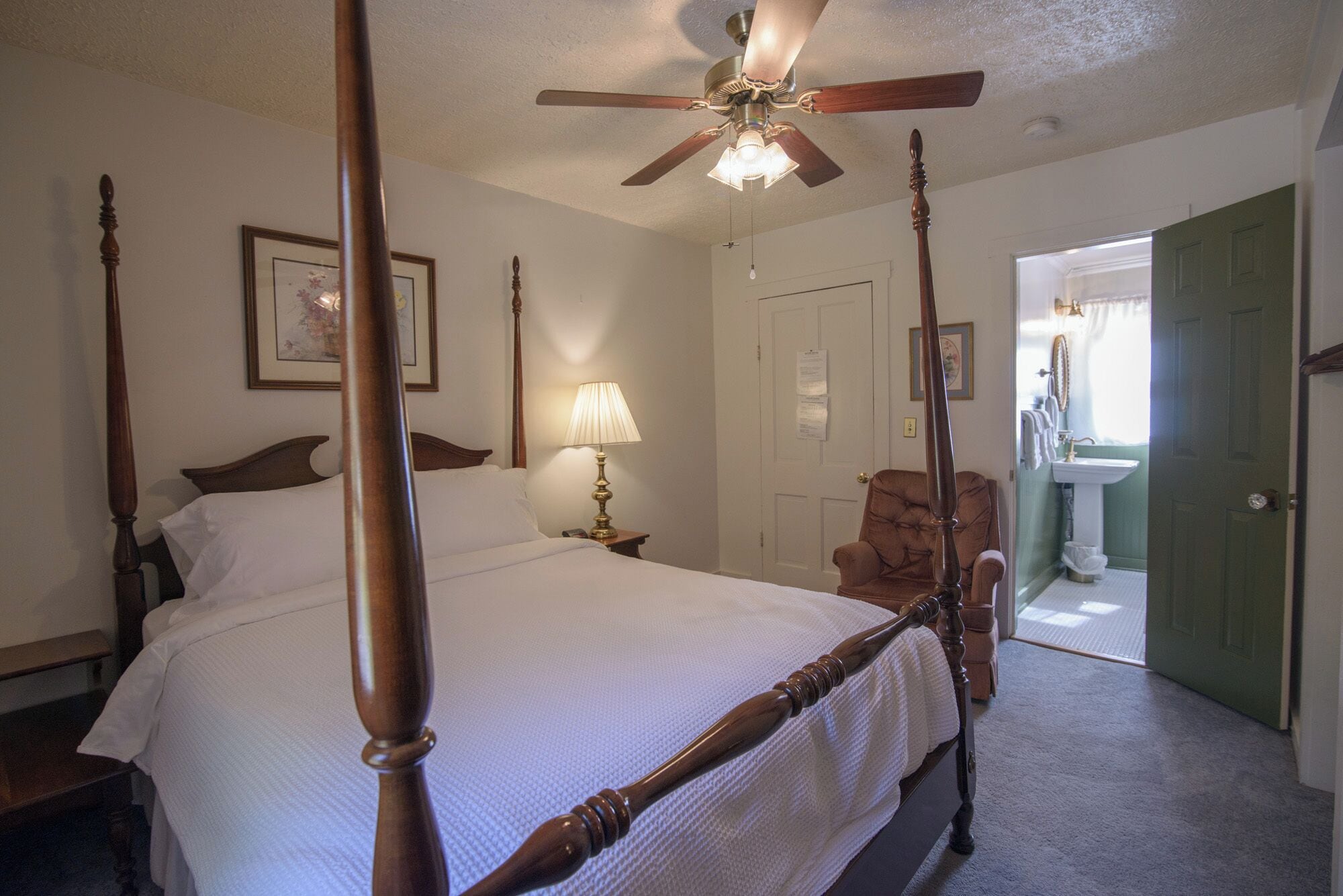 A four-poster bed with a ceiling fan, a bedside lamp, and a chair in a room with a door leading to a bathroom.