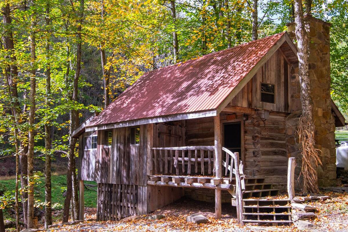 A rustic cabin with a wooden porch and a stone wall, surrounded by autumn foliage.