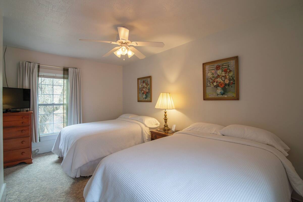 A hotel room with two beds, a ceiling fan, a TV, and two paintings on the wall.