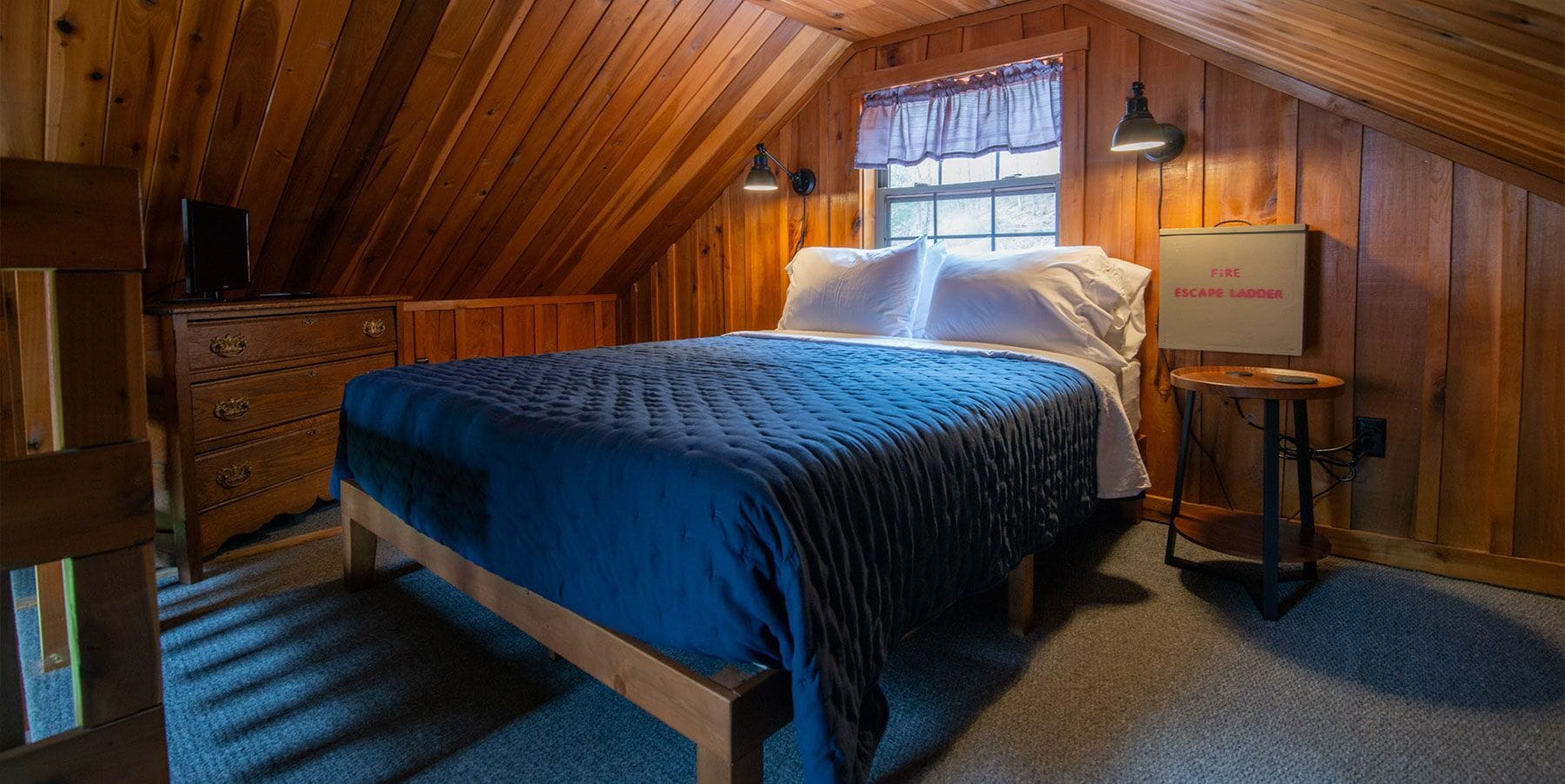 A cozy attic bedroom with a wooden sloped ceiling, a bed with a blue quilt, a dresser, a nightstand, and a fire escape ladder.