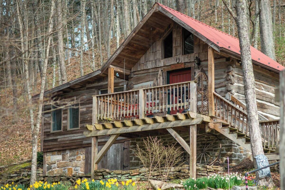 A rustic log cabin with a wooden staircase leading to a balcony, surrounded by a stone wall and a garden with daffodils.