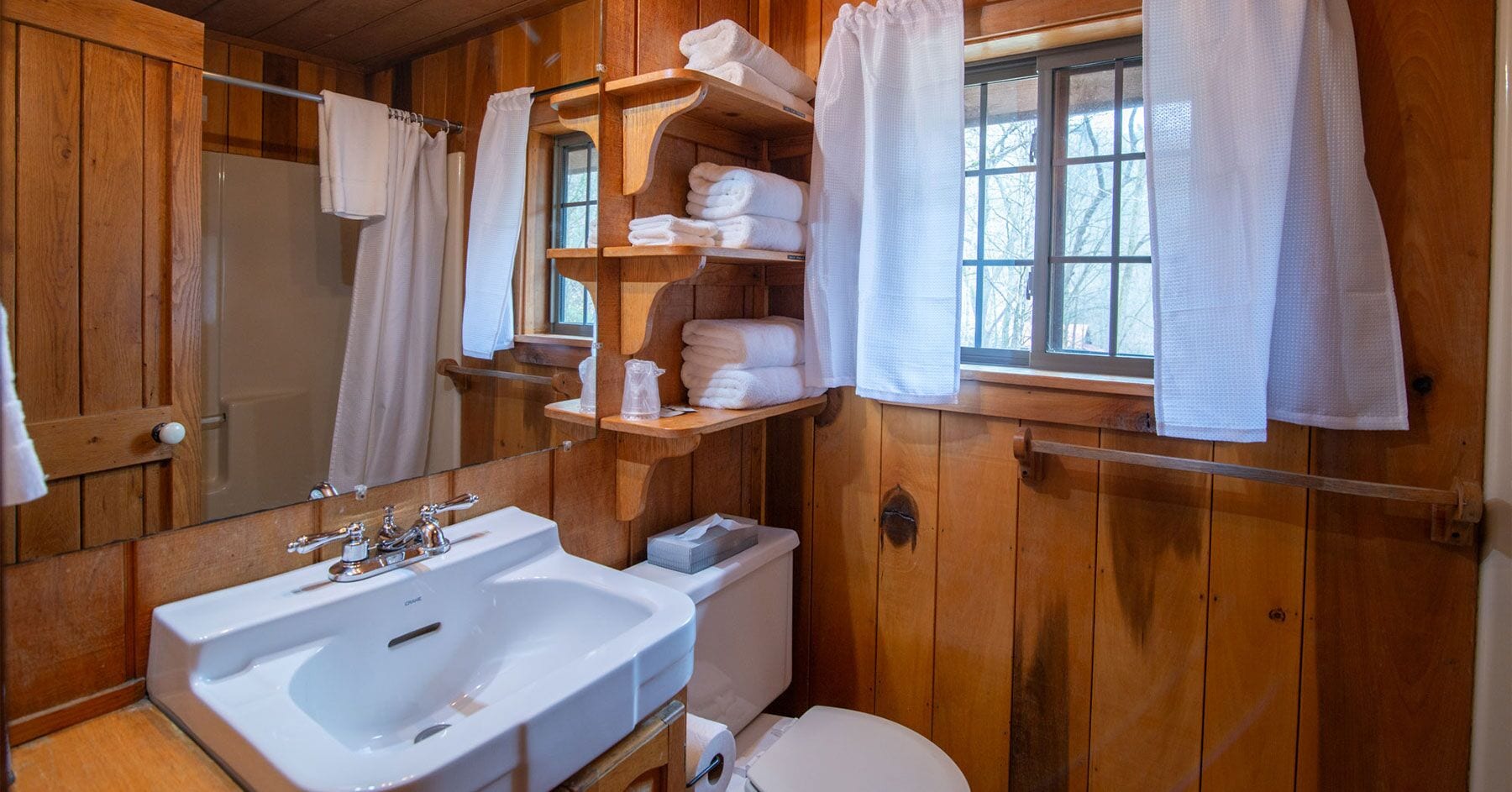 A wooden cabin bathroom with a sink, toilet, and shelves holding towels.