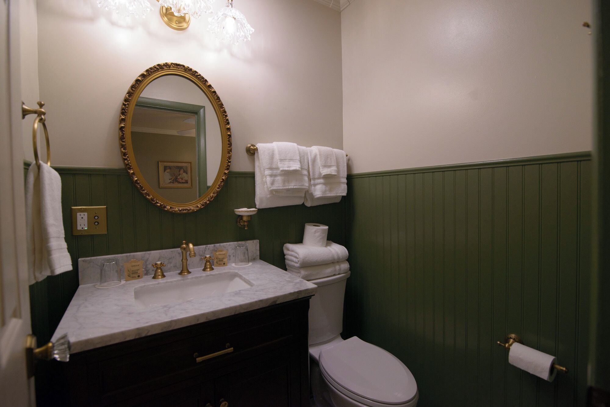 A bathroom with a marble sink, gold faucets, a white toilet, and green wainscoting.