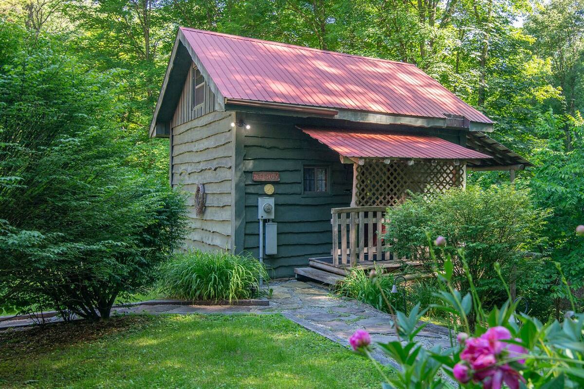A rustic cabin with a red roof, surrounded by lush greenery and a stone pathway leading to the entrance.