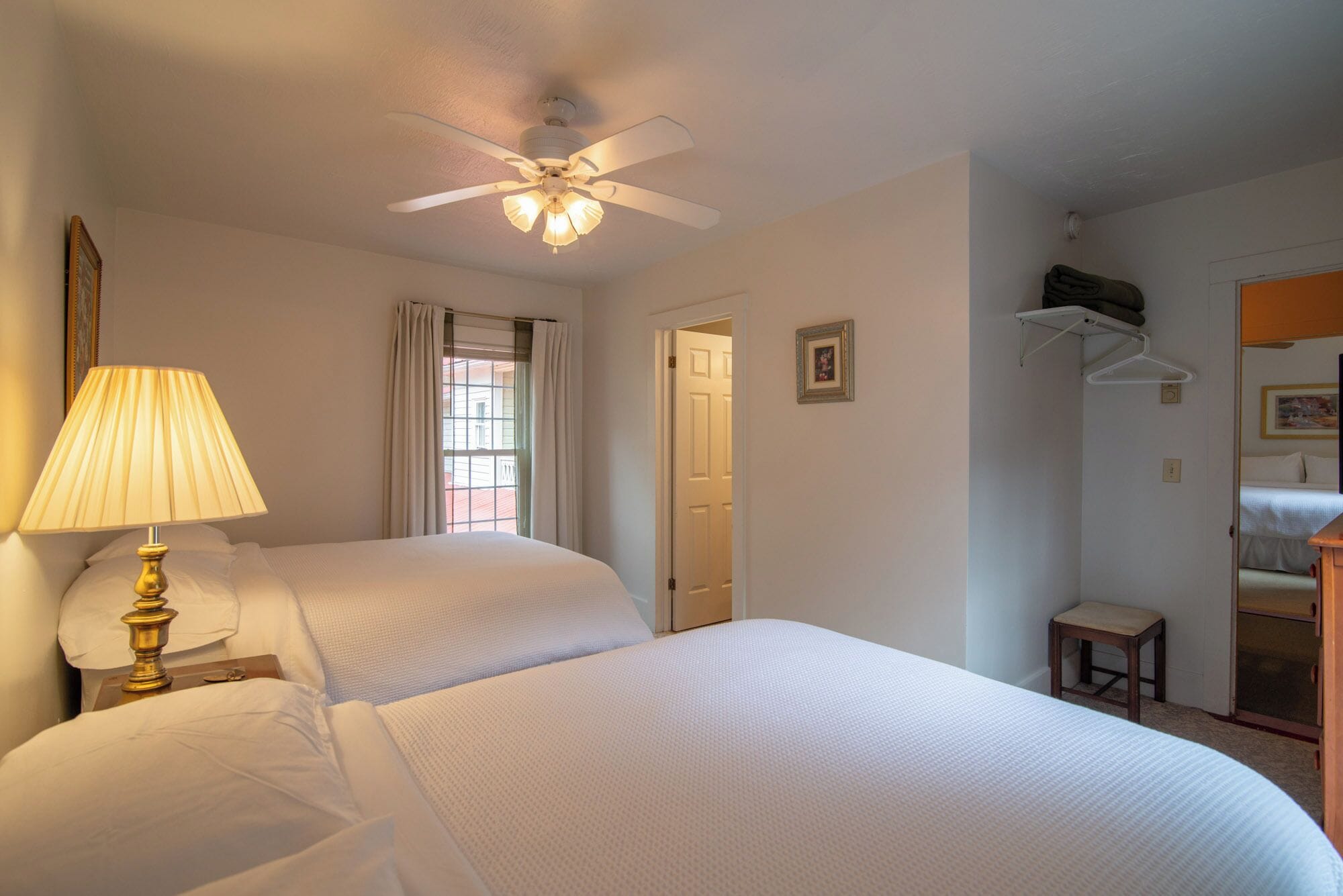 Une chambre d’hôtel avec deux lits, un ventilateur de plafond, une lampe, une fenêtre avec des rideaux, une porte, une petite table et un porte-manteau.