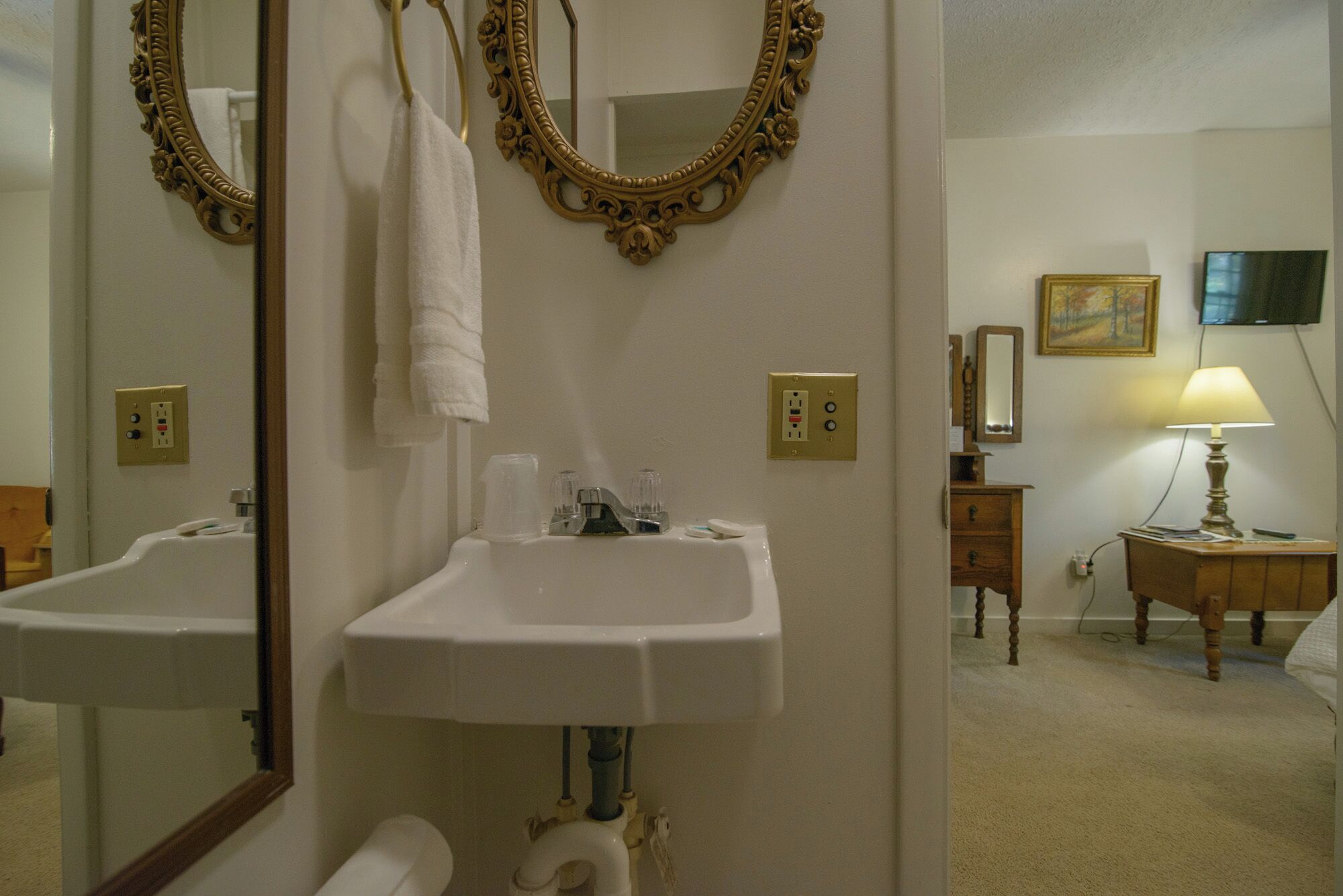 A bathroom with two sinks, a mirror, a towel hanging on the wall, a painting, and a lamp on a bedside table.