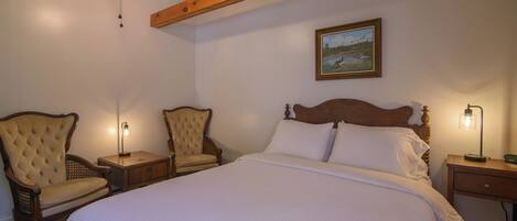 A bedroom with a large bed, two upholstered chairs, a ceiling fan, and a painting on the wall.