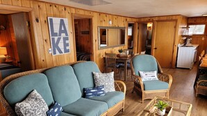 Ein gemütliches Blockhaus-Interieur mit einem grünen Sofa, Korbstühlen, einem kleinen Tisch mit einer Pflanze und einer Küchenzeile im Hintergrund.