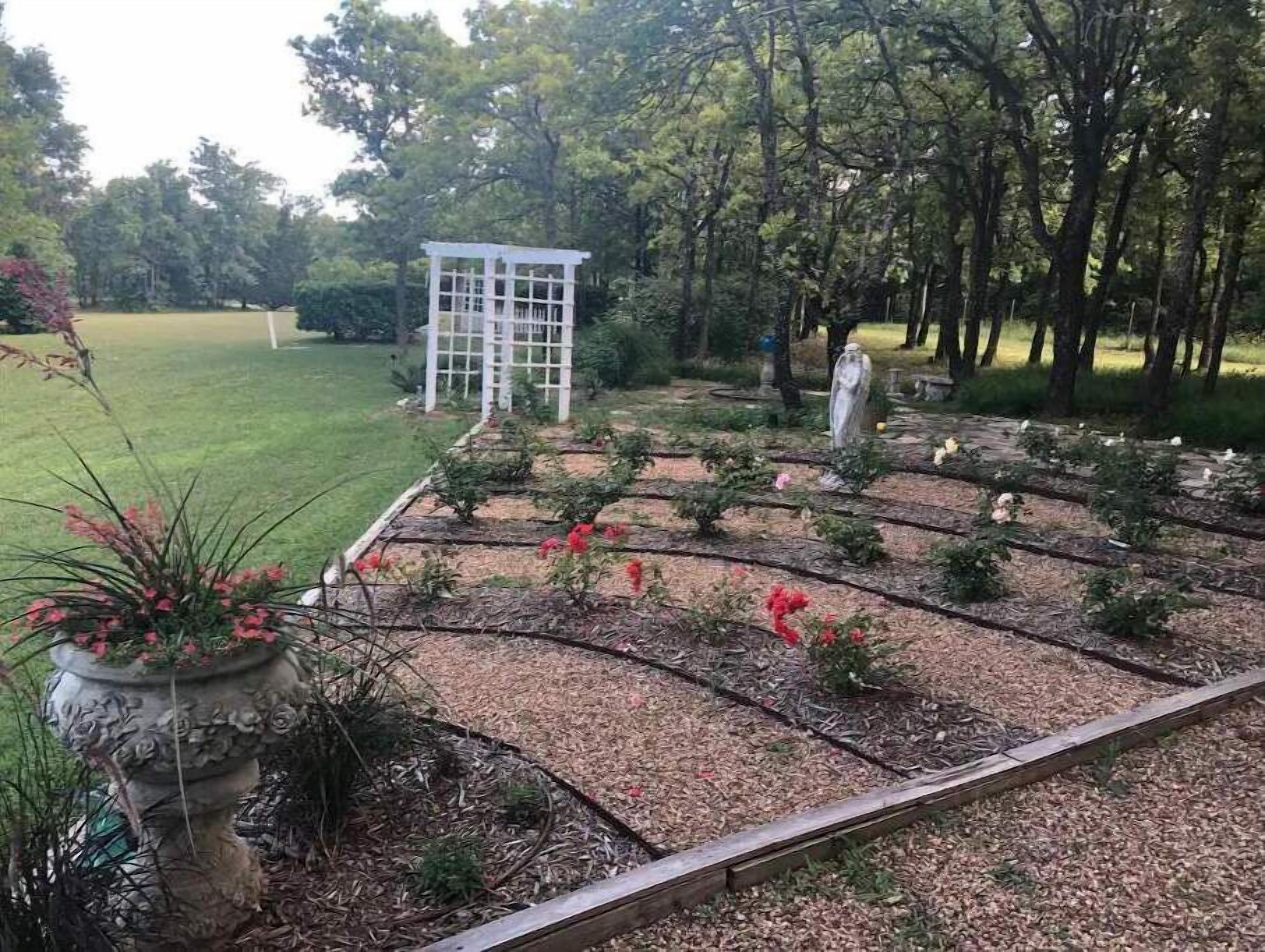 Ein Garten mit einer weißen Pergola, einer Statue und einem Blumenbeet mit roten Blüten.