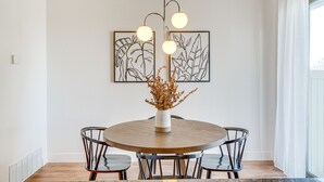 A dining area with a round wooden table, four chairs, a vase with dried flowers, and two framed artworks on the wall.