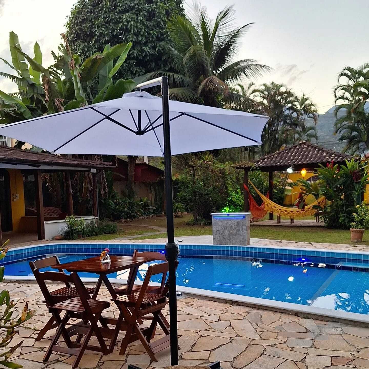 Un espace piscine aménagé avec une table et des chaises, entouré de plantes tropicales et agrémenté d’un hamac.