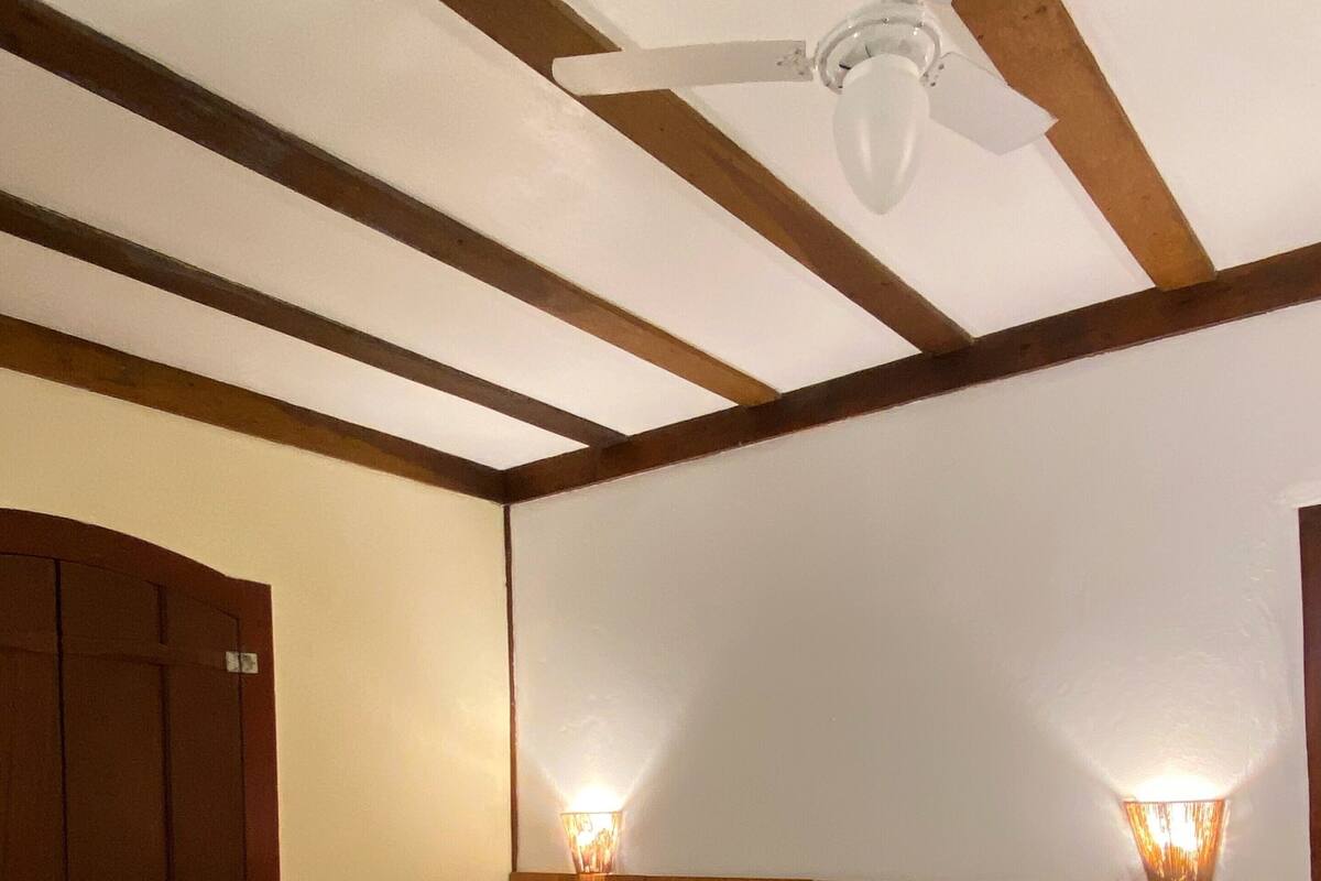 A hotel room with a bed, a ceiling fan, wooden beams, a nightstand, and two wall-mounted lights.