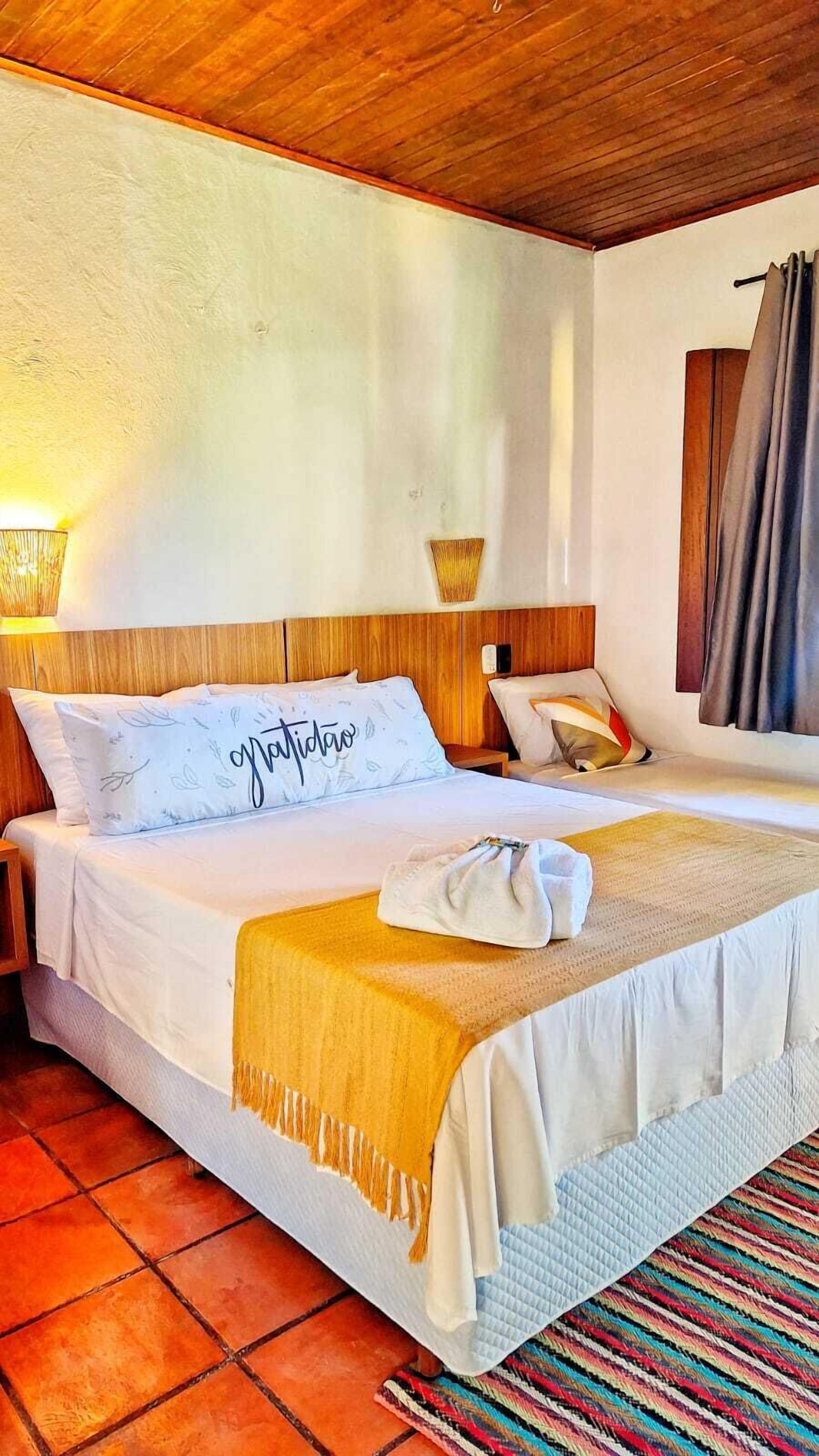 Een net opgemaakt bed met een witte deken en een kussen met de tekst 'gratitude', een houten hoofdbord, een bedlampje en een gestreept vloerkleed.