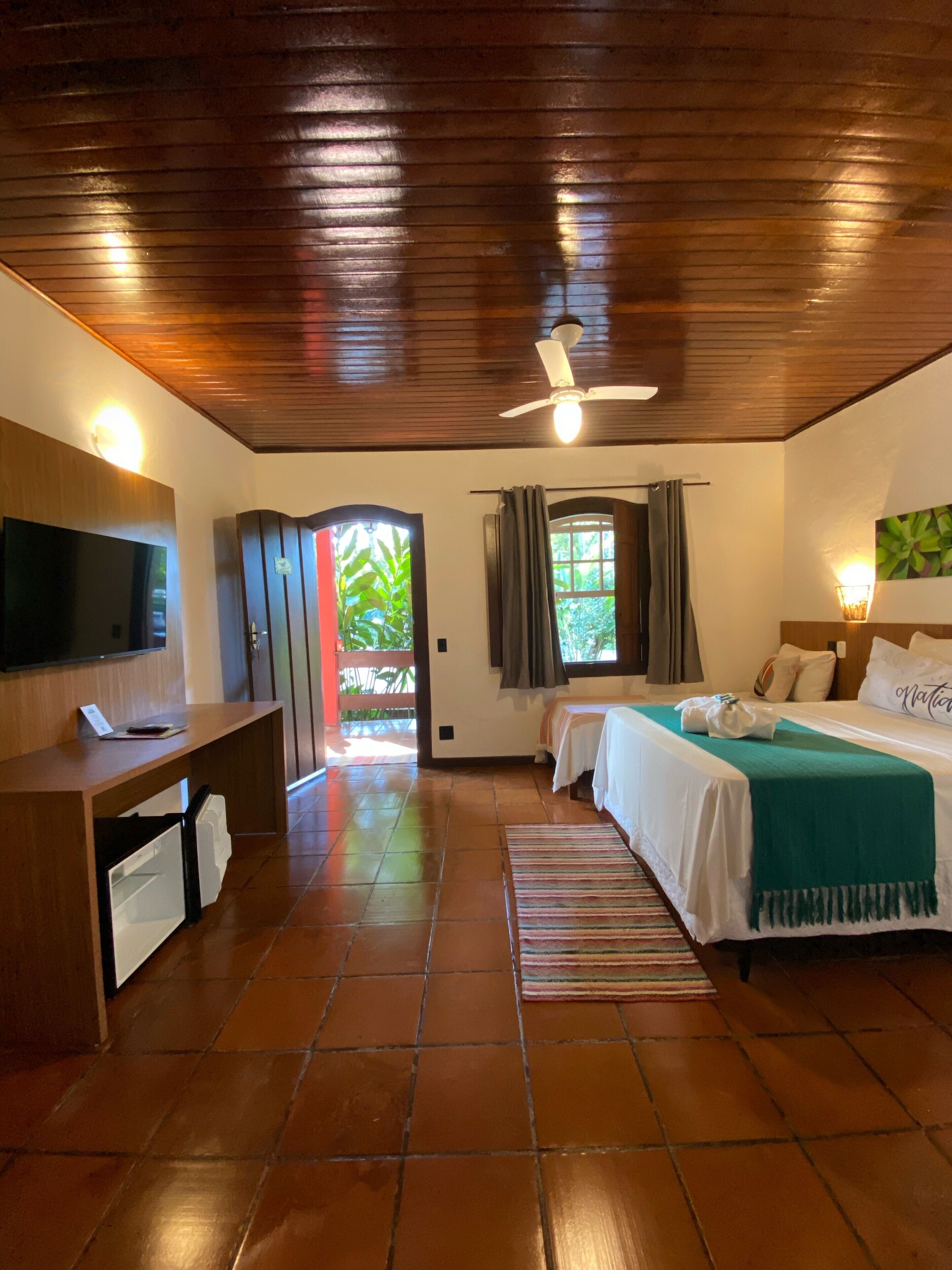 A hotel room with a wooden ceiling, a bed with a green blanket, a desk with a TV, and a window with curtains.