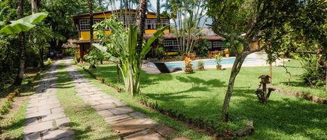 Un sendero conduce a un edificio rodeado de abundante vegetación y una piscina.