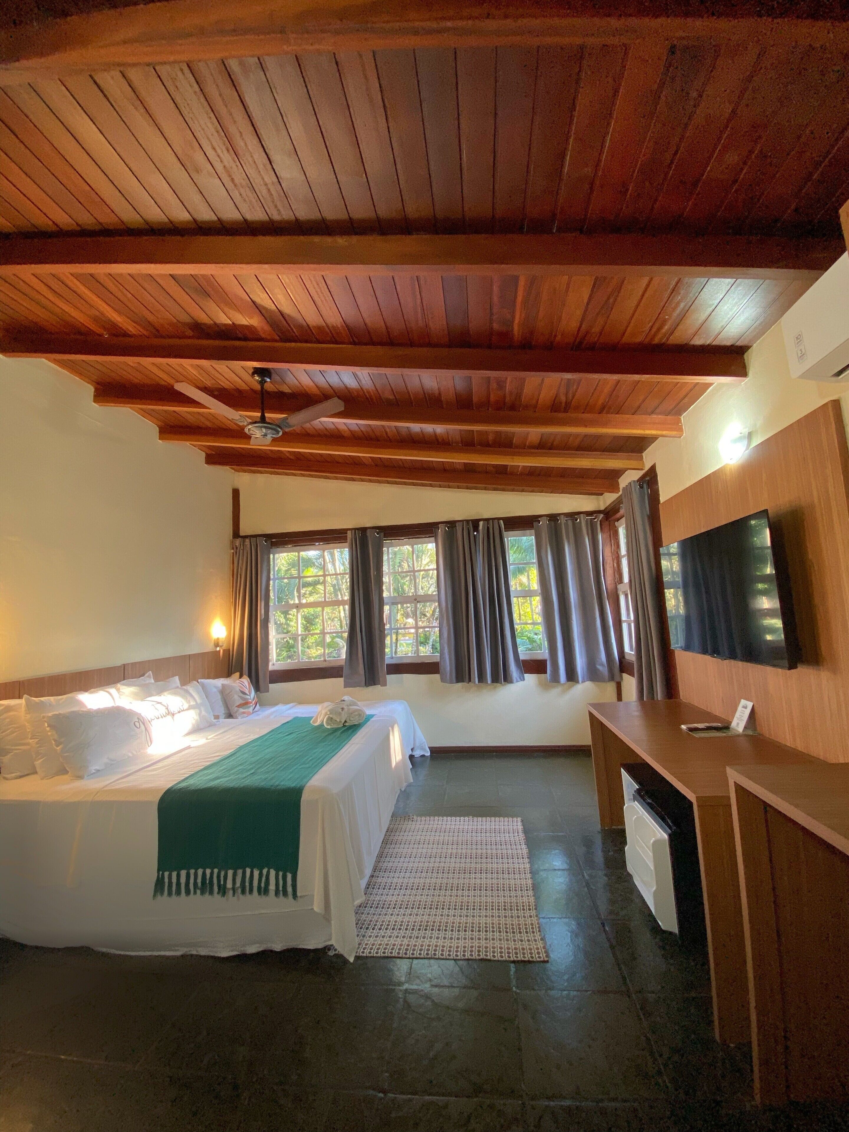 A hotel room with a wooden ceiling, a bed with white bedding, a desk, and a TV.