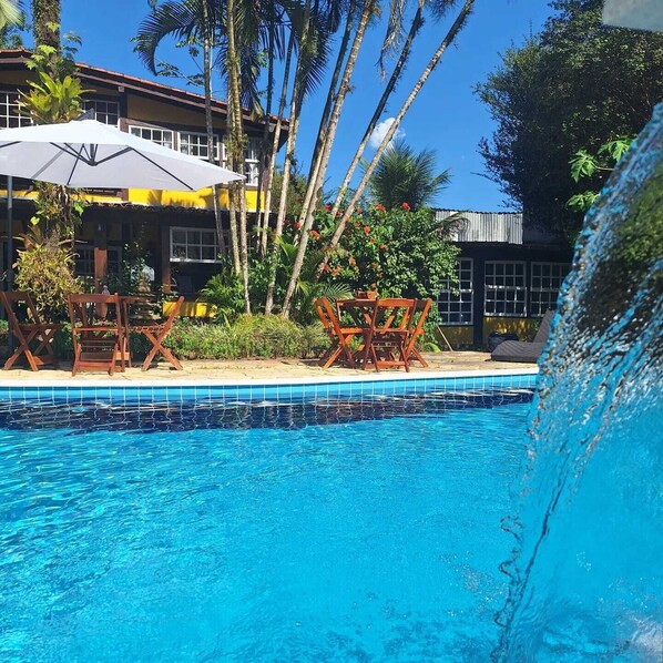 Una piscina con agua azul cristalina, rodeada de sillones de madera y sombrillas, con palmeras y un edificio al fondo.