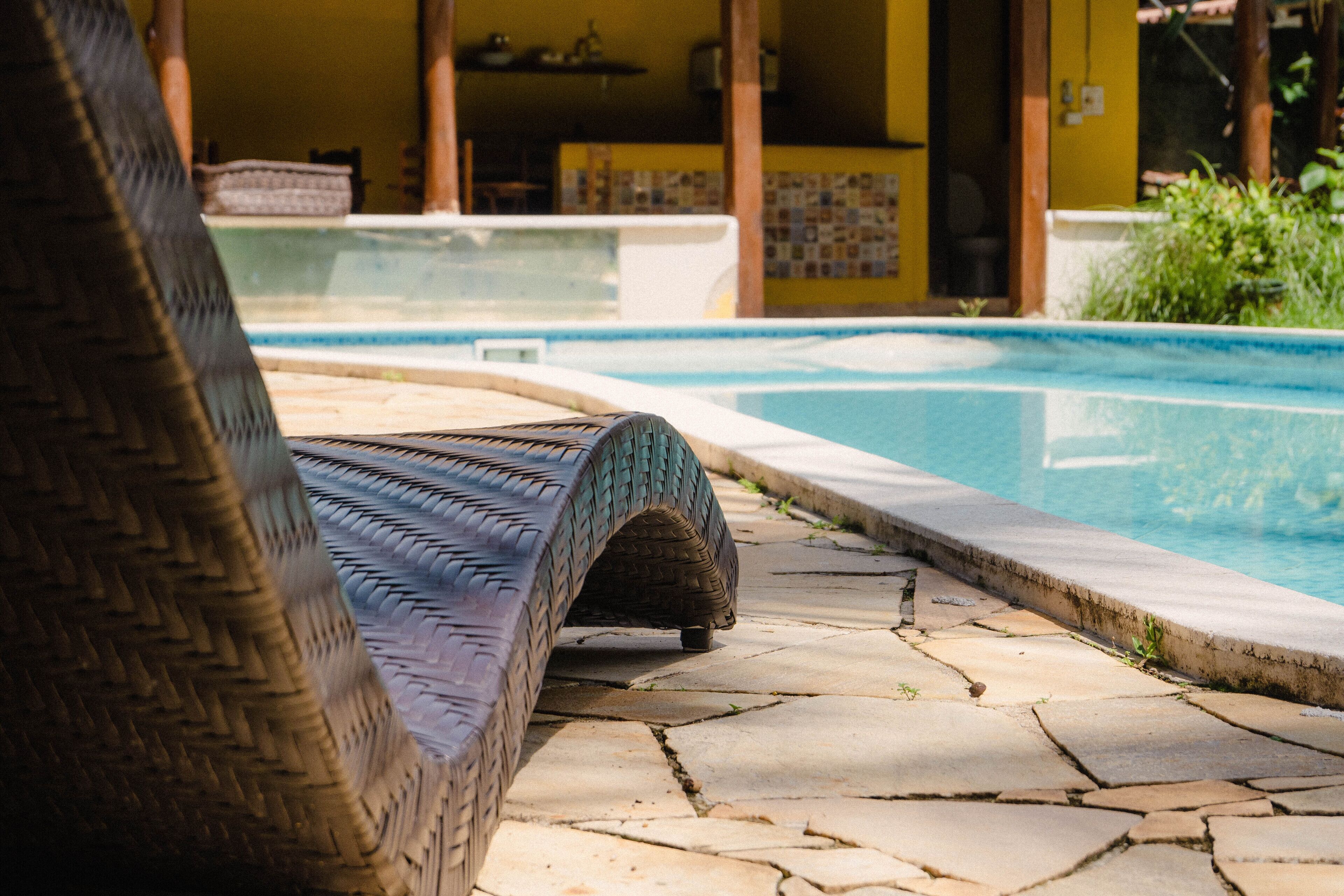 Una silla de mimbre junto a una piscina con baldosas de piedra y una pared amarilla al fondo.