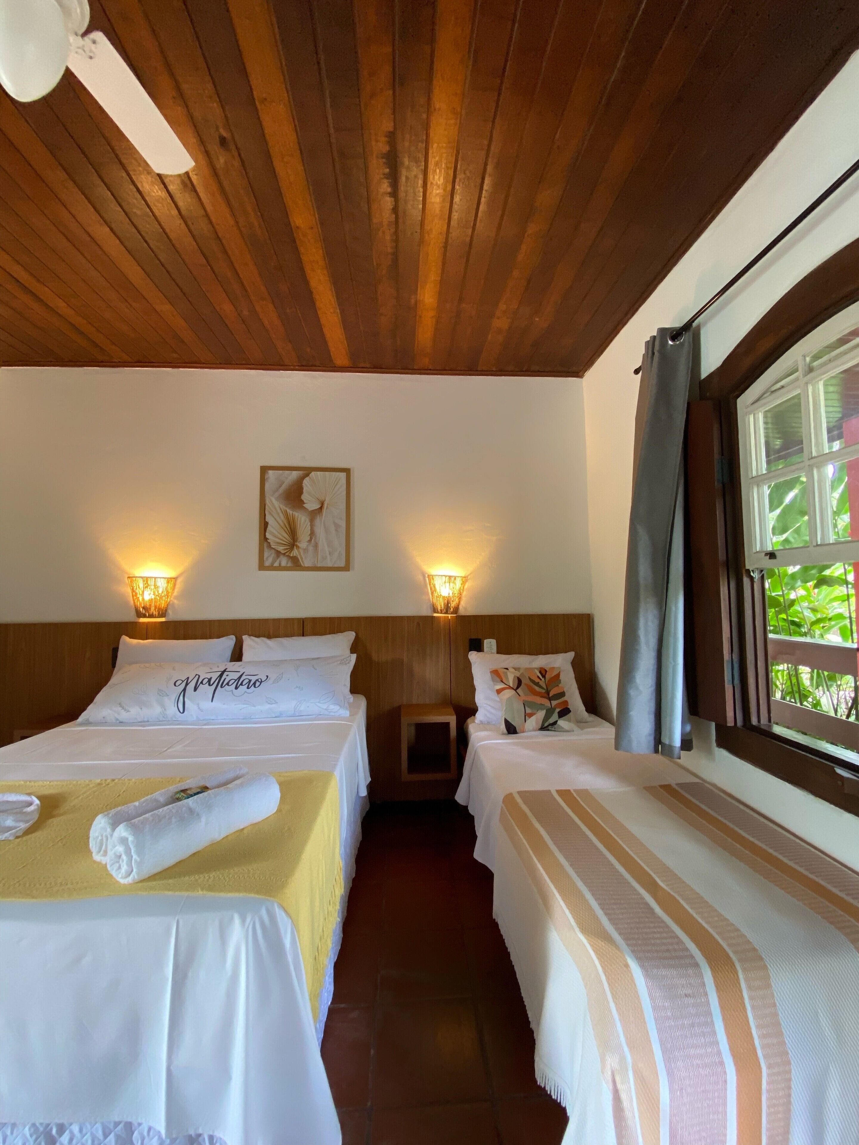 Een hotelkamer met twee bedden, een houten plafond, wandlampen, een raam met gordijnen, en een klein nachtkastje.