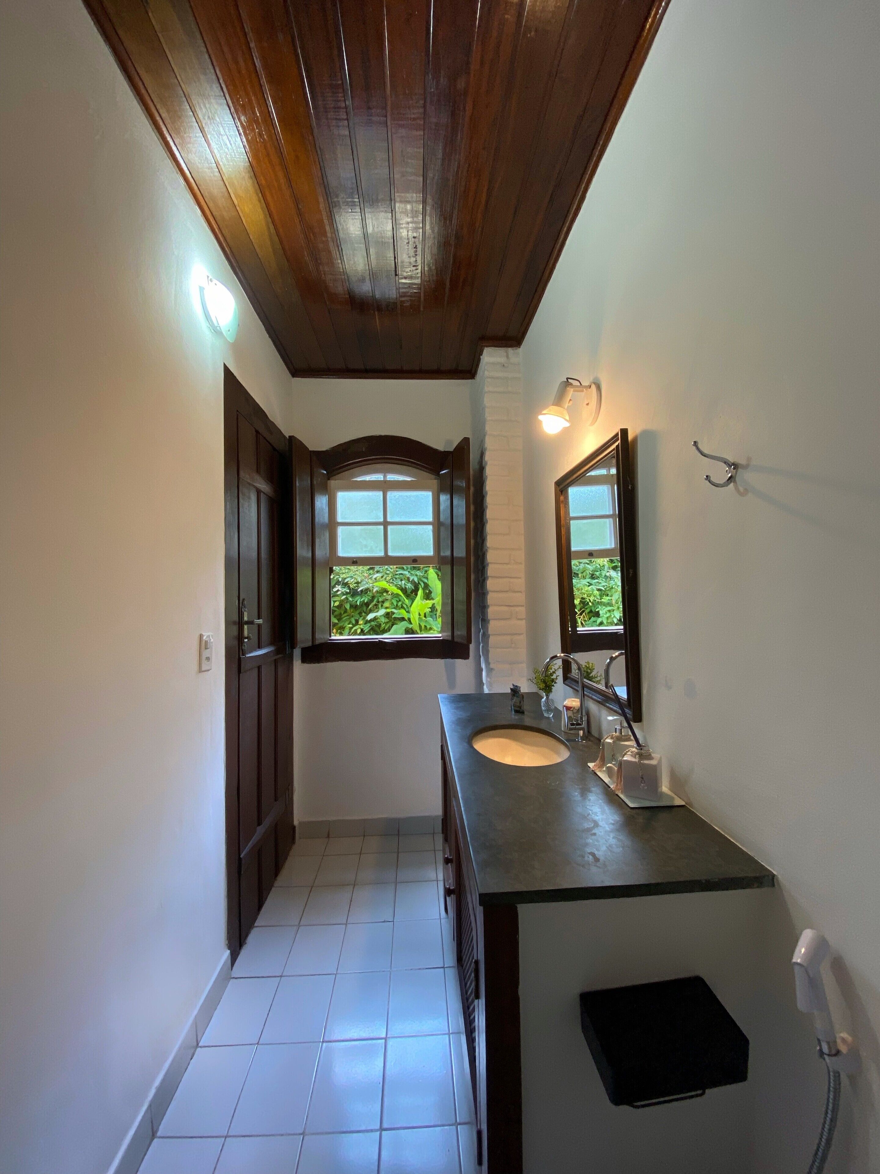 A narrow bathroom with a wooden ceiling, a window with a view of greenery, a sink with a mirror, and a shower area.
