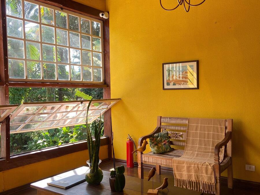 Una habitación con techo de madera, un ventanal con vistas a la vegetación, una mesa de madera con un florero y una figurita de pájaro, y un banco con cojín.