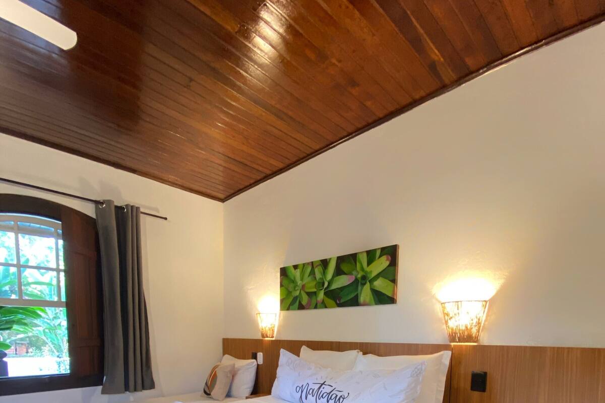 A hotel room with two beds, a green throw blanket, a wall-mounted light, and a window with curtains.