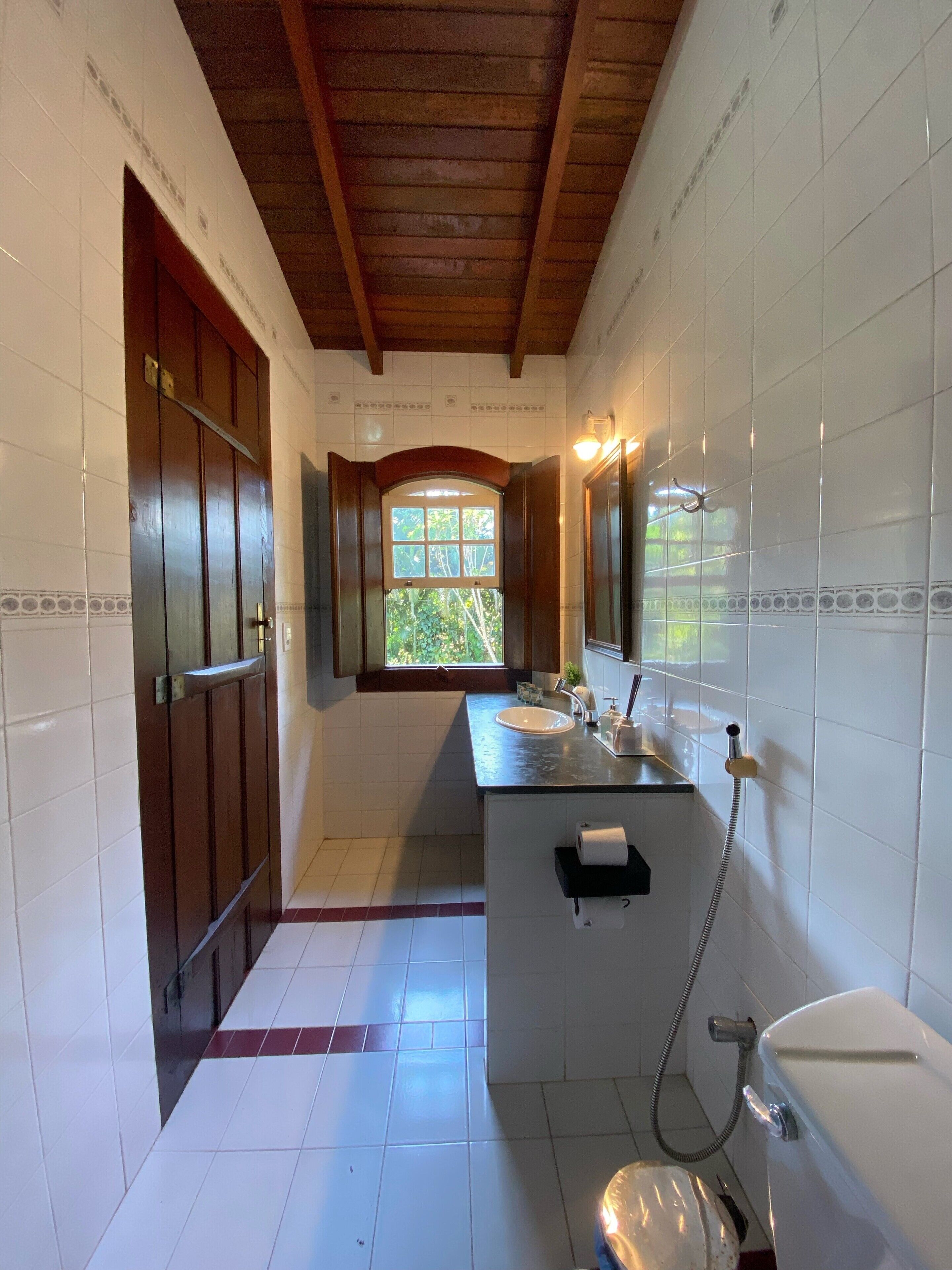A bathroom with a wooden ceiling, white tiled walls, a window, a sink, and a toilet.