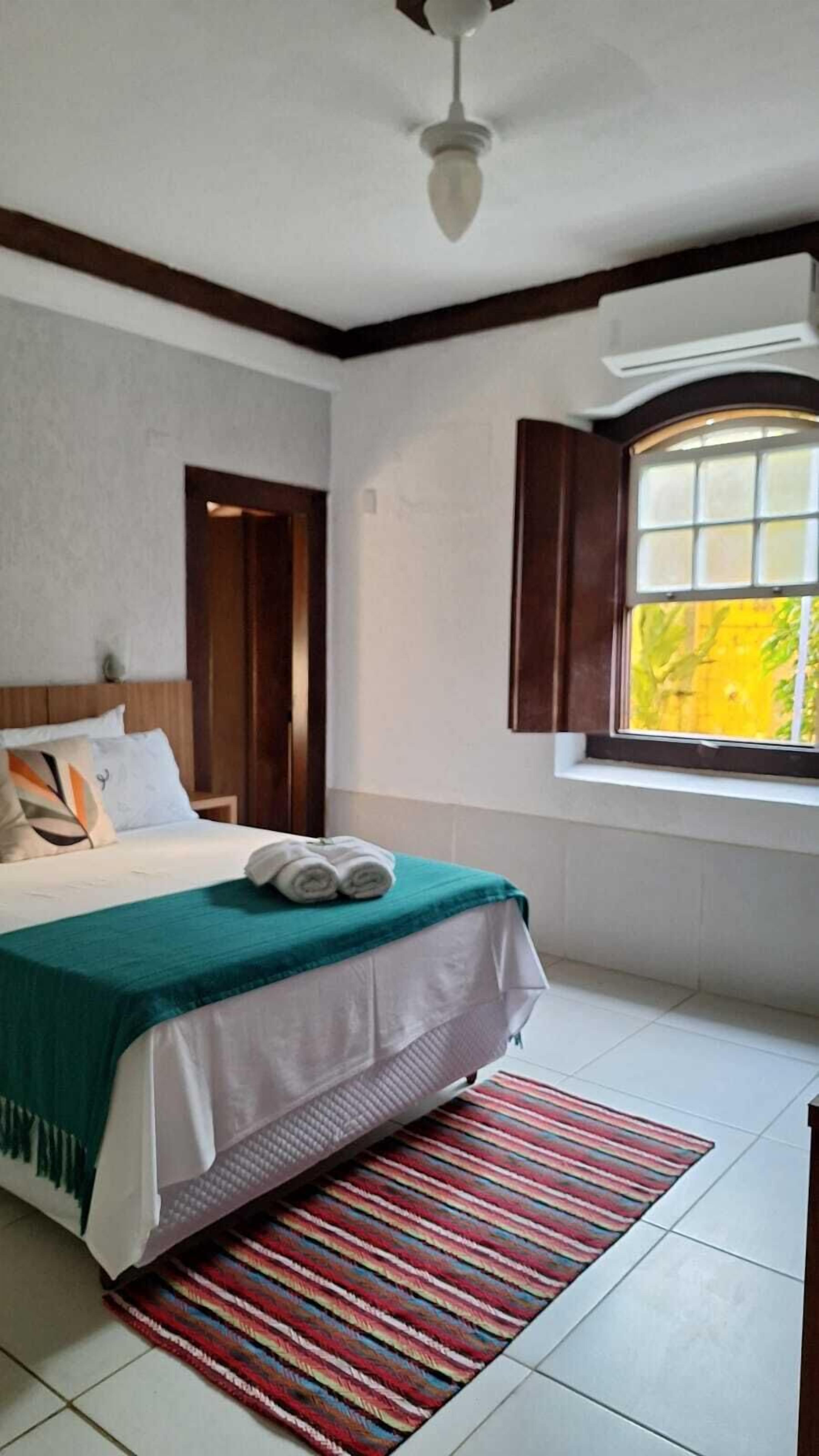 Une chambre avec un lit, une fenêtre avec des rideaux, un ventilateur de plafond et une vue sur la verdure à l’extérieur.