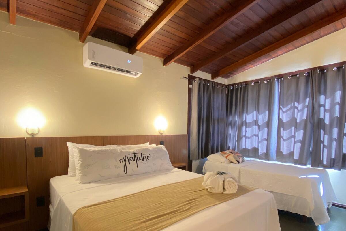A hotel room with a wooden ceiling, a bed with a white bedspread and a gold throw blanket, a bedside table with a lamp, and a window with curtains.