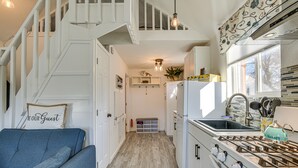 Cucina compatta con piano cottura a gas, mobili bianchi e forno integrato. C'è un divano blu con un cuscino, una piccola mensola con una statuetta di un cane e una finestra vista alberi.