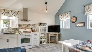 Una cucina moderna con mobili bianchi, un forno a microonde integrato e un piano cottura a gas. C'è un'area pranzo con tavolo e sedie bianche, e un piccolo tavolino in legno con un televisore.