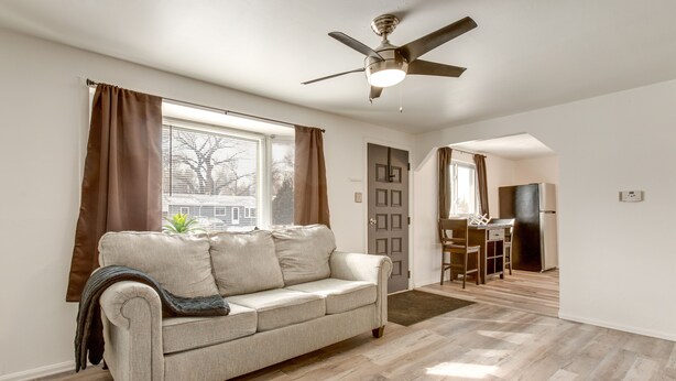 A living room with a beige sofa, a ceiling fan, and a view of a cityscape through the window. - Covered Patio, Gas Grill & Yard: Sheridan Home! (Sheridan)
