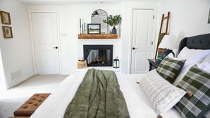 A bedroom with a fireplace, a bed with green and white bedding, a nightstand with a lamp, and a wooden shelf above the fireplace. - New Year’s Retreat. Cozy Modern Farmhouse! Close to historic downtown Wylie! (Wylie)