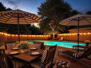 A poolside area with a dining table set for two, surrounded by chairs, and two large umbrellas providing shade.