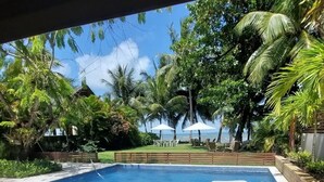 A swimming pool with white lounge chairs, surrounded by lush greenery and palm trees, with a view of the ocean. - Casa Amendoeiras Charme e Conforto à Beira-mar (Ponta de Serrambi Ipojuca)
