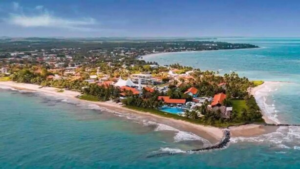 An aerial view of a coastal resort with a beach, a swimming pool, and a large building with a red roof. - Casa Amendoeiras Charme e Conforto à Beira-mar (Ponta de Serrambi Ipojuca)