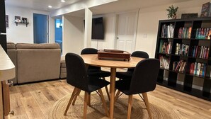 Un coin repas avec une table ronde en bois, des chaises noires et un tapis tissé. Une bibliothèque remplie de livres et une plante se trouvent à droite. Un canapé et un téléviseur sont visibles au fond.