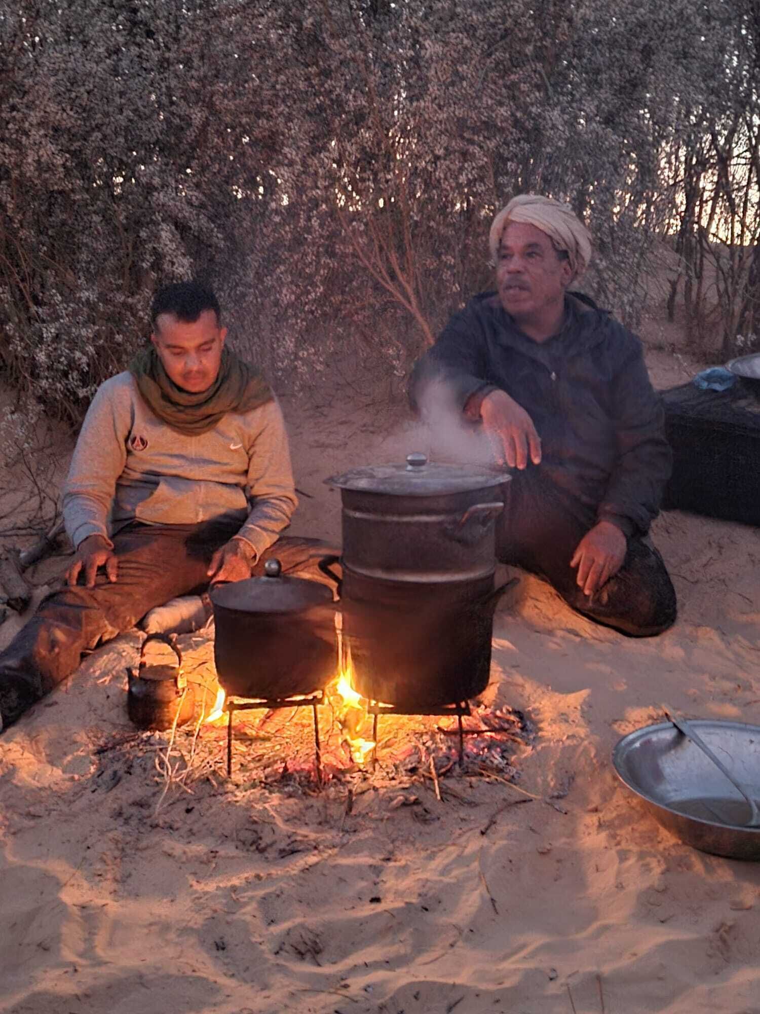 Deux personnes sont assises près d’un feu de camp, avec des marmites pour cuisiner, dans un environnement extérieur sablonneux.