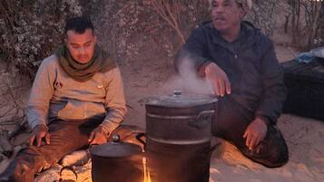 Deux personnes sont assises près d’un feu de camp, avec des marmites pour cuisiner, dans un environnement extérieur sablonneux.