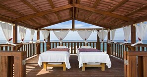 Une terrasse en bois avec deux lits, des rideaux blancs et une vue sur la mer.