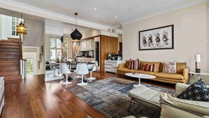 A spacious living room with a wooden floor, a brown sofa, white armchairs, and a coffee table. There is a kitchen in the background with modern appliances and a dining area with white chairs. - Chic Chelsea maisonette with 2 terraces (London)